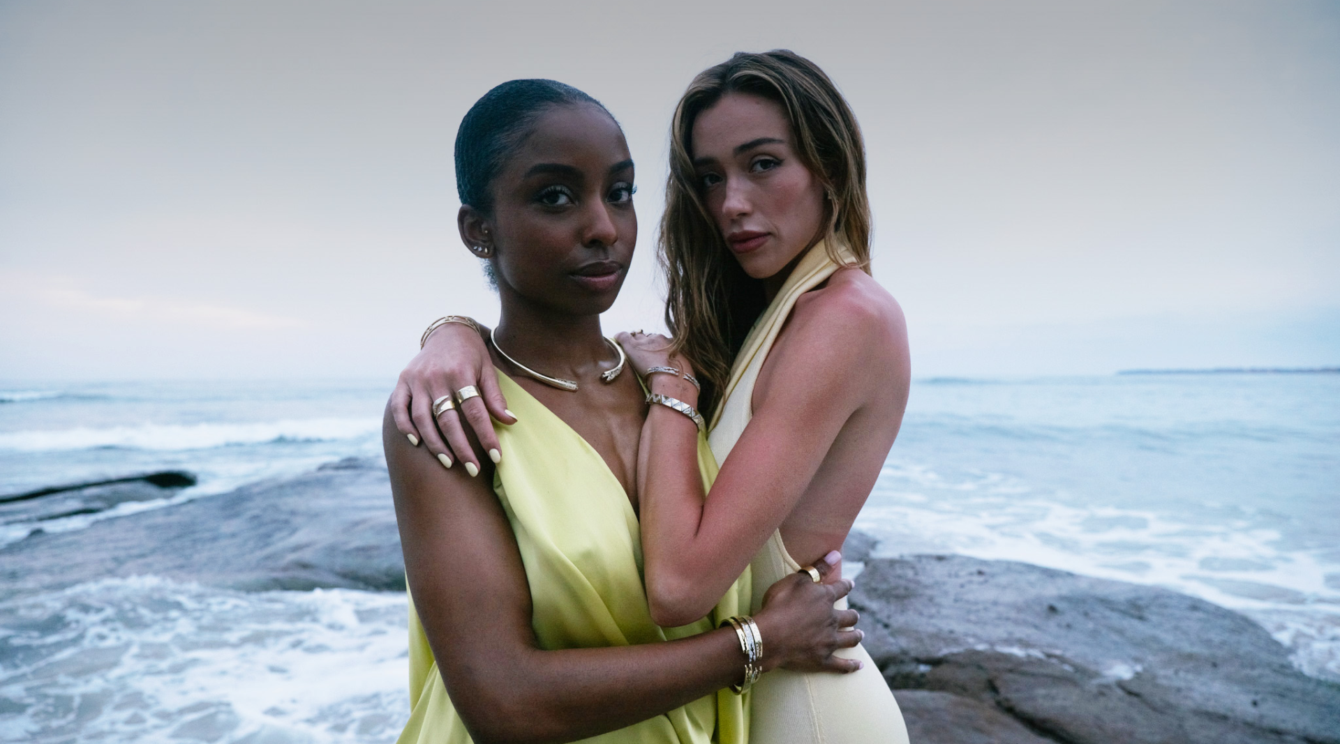 Two women in yellow dresses standing on a rocky beach with ocean waves in the background.