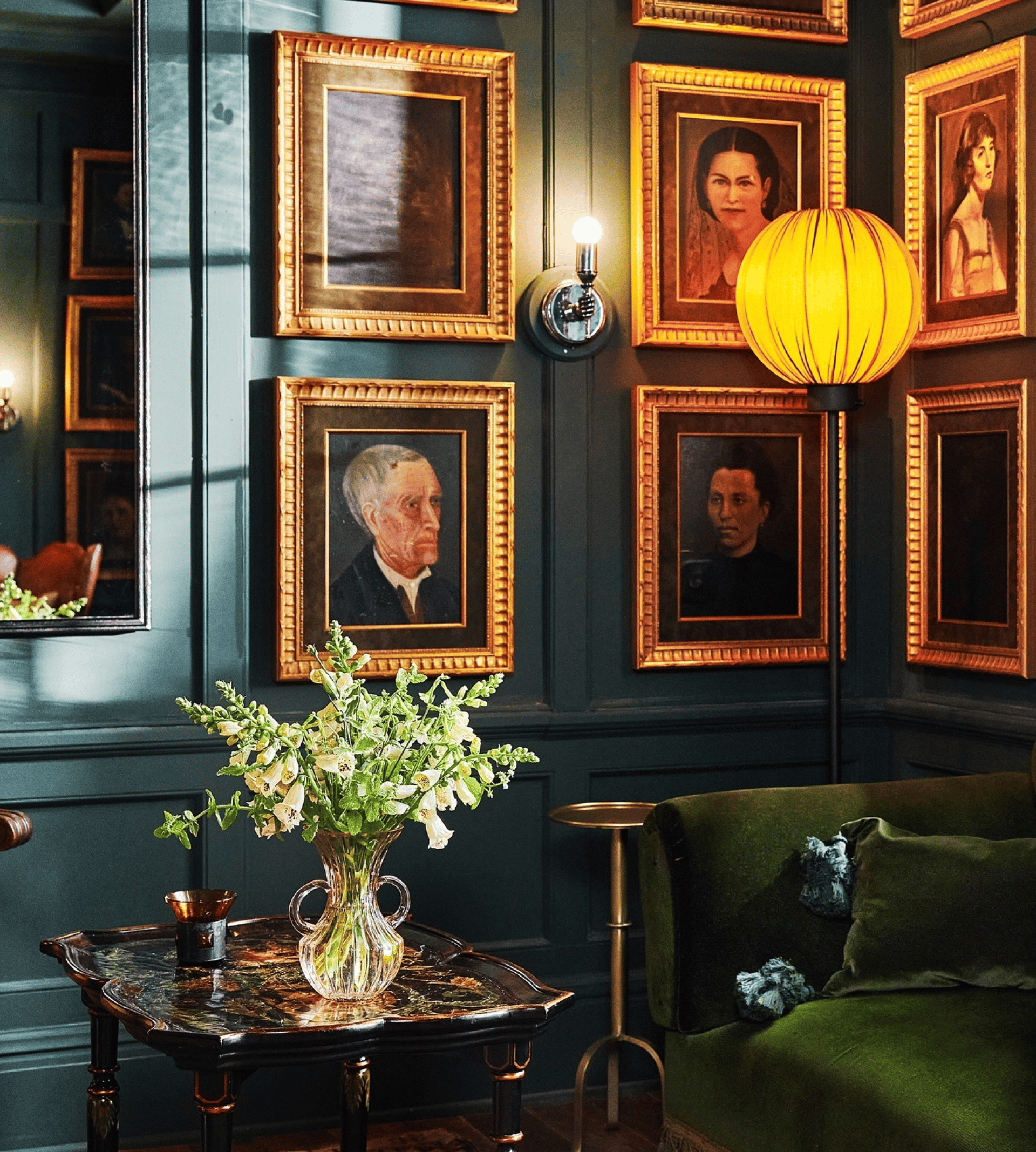 Decorative interior with framed portraits on a dark wall, a yellow pendant light, and a green sofa.