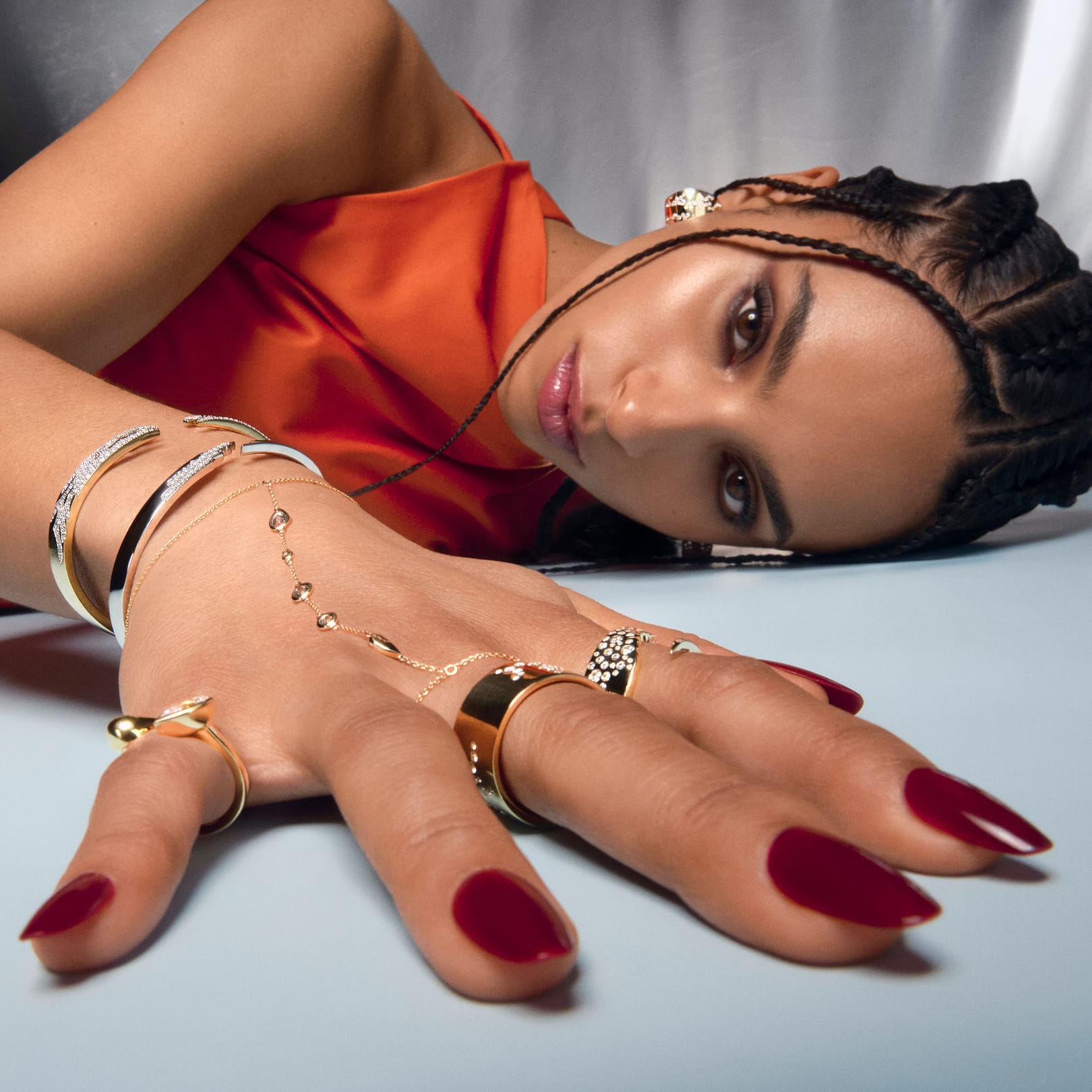 A person with red nails lying down, showcasing gold hand chain jewelry on their hands.