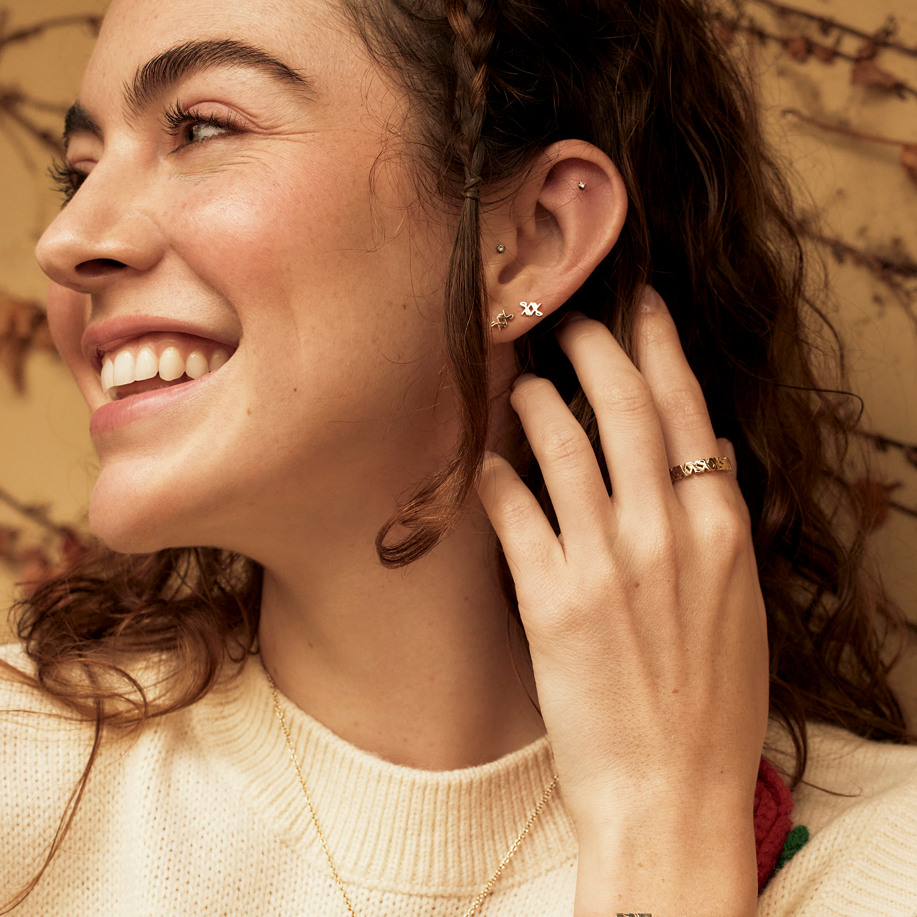 Model wearing 14k yellow gold SAV logo stud earrings as part of a layered ear stack, paired with a SAV eternity ring and a delicate chain necklace, in a warm outdoor editorial setting.
