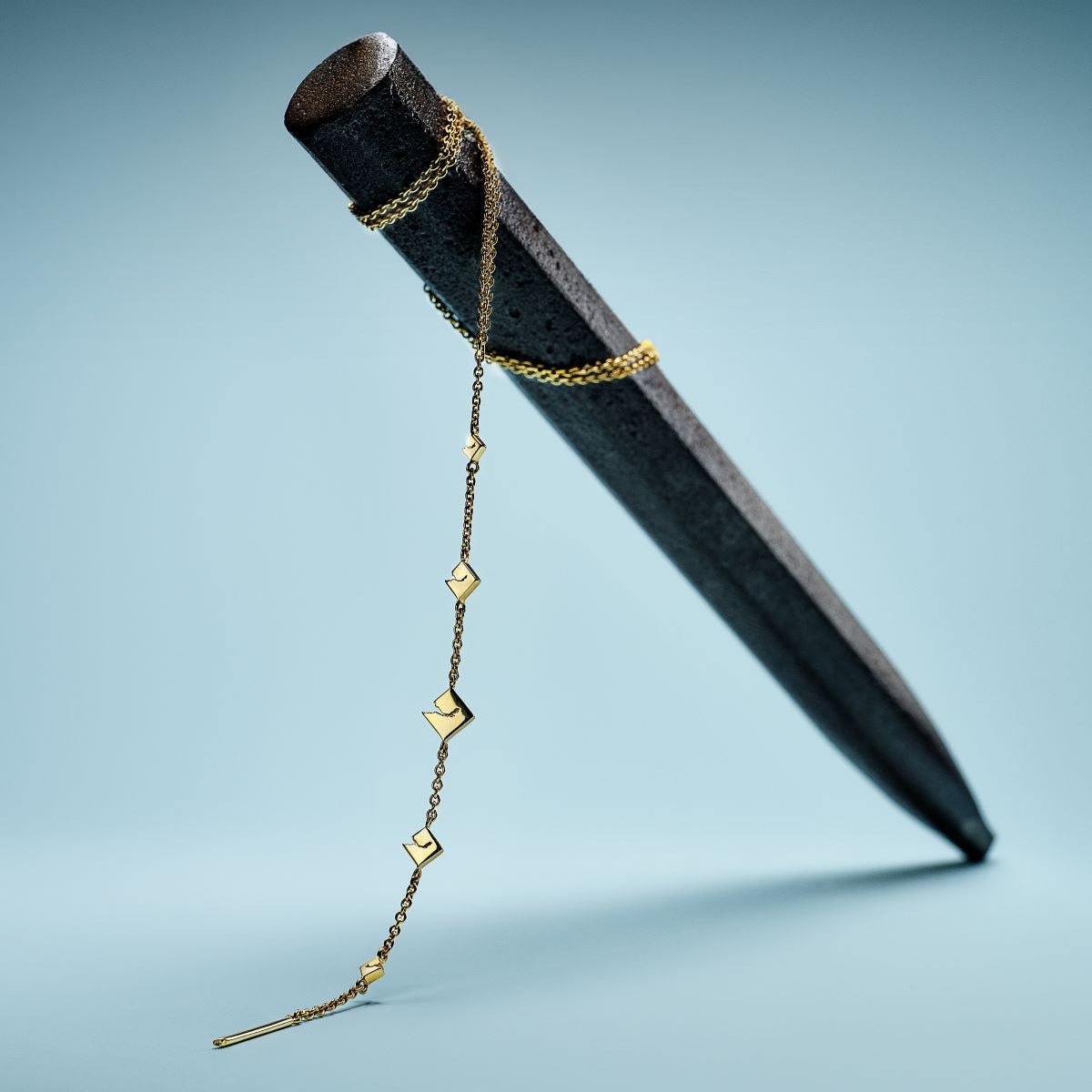 The yellow gold version of the Y-lariat necklace draped and hanging from a dark weathered metal cylindrical prop, the geometric diamond-shaped station links and slim bar pendant visible against a teal background.