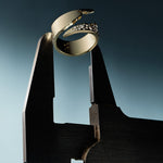 A gold bypass ring with diamond accents is displayed on a stand against a gradient blue background.