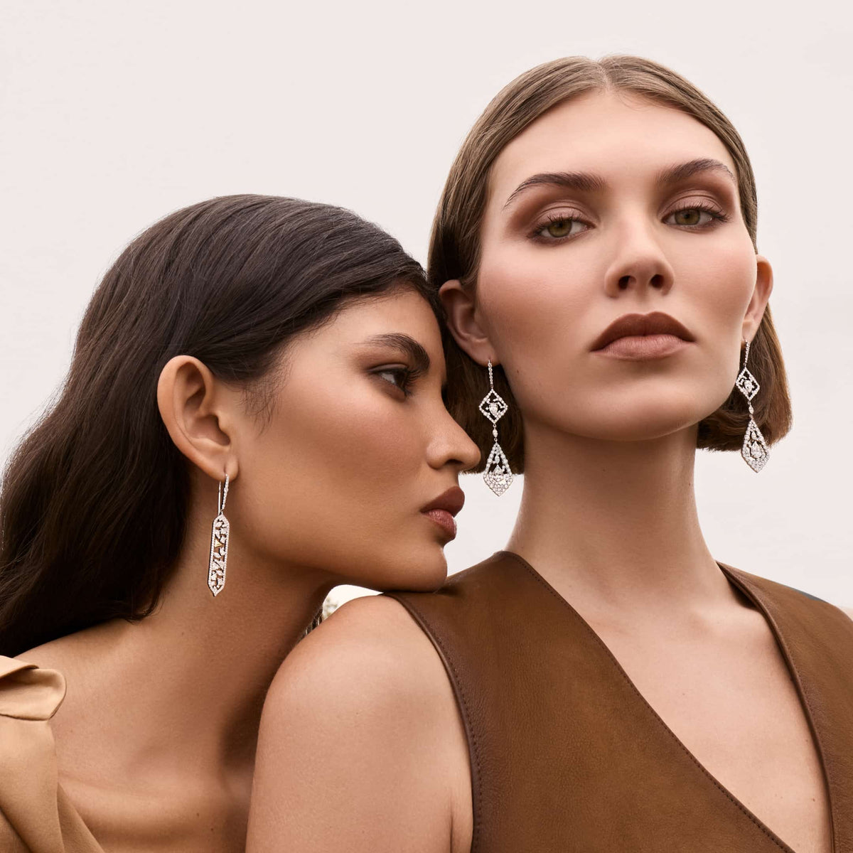 Two models facing forward against a soft neutral background, the model on the left wearing the 14k white gold mixed fancy-cut diamond drop earring, and the model on the right wearing a different multi-drop chandelier-style diamond earring, both in brown tonal outfits.