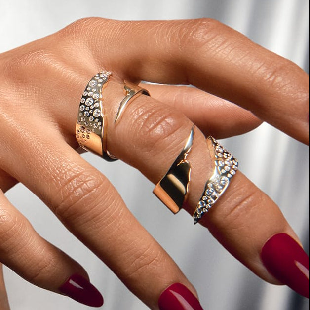A model wearing a stack of 14k yellow gold SAV collection rings across multiple fingers and a layered cuff bracelet stack on her wrist, extending her hand toward the camera against a silver draped backdrop