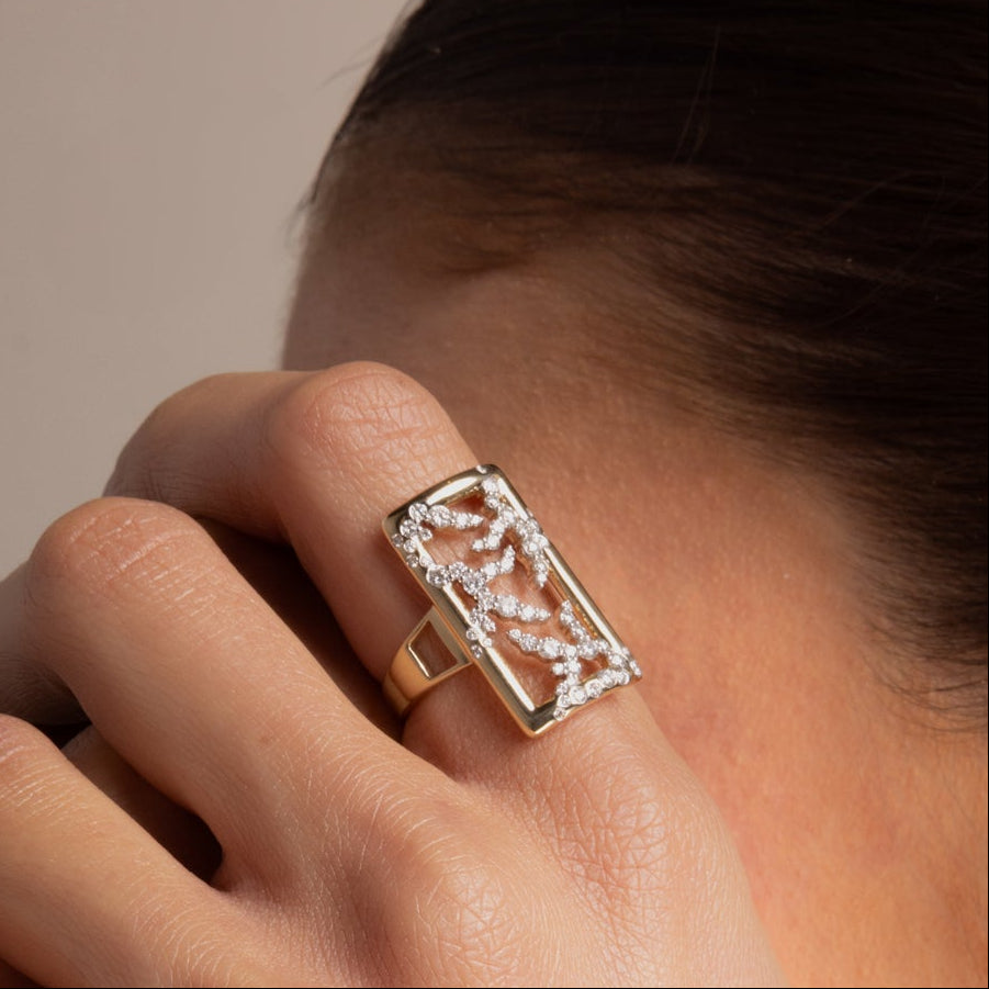 A model wearing the white gold rectangle SAV diamond logo cocktail ring on her index finger and the yellow gold rectangle SAV diamond logo hoop earring in her ear, with her hand raised near the side of her face against a warm taupe backdrop.