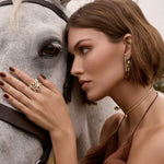 Close-up profile of a model pressing her cheek against a gray horse, wearing the two halves of the 14k yellow gold puzzle ring separated and stacked as individual rings on her index finger, styled alongside the rectangle SAV logo hoop earrings and a gold collar necklace.
