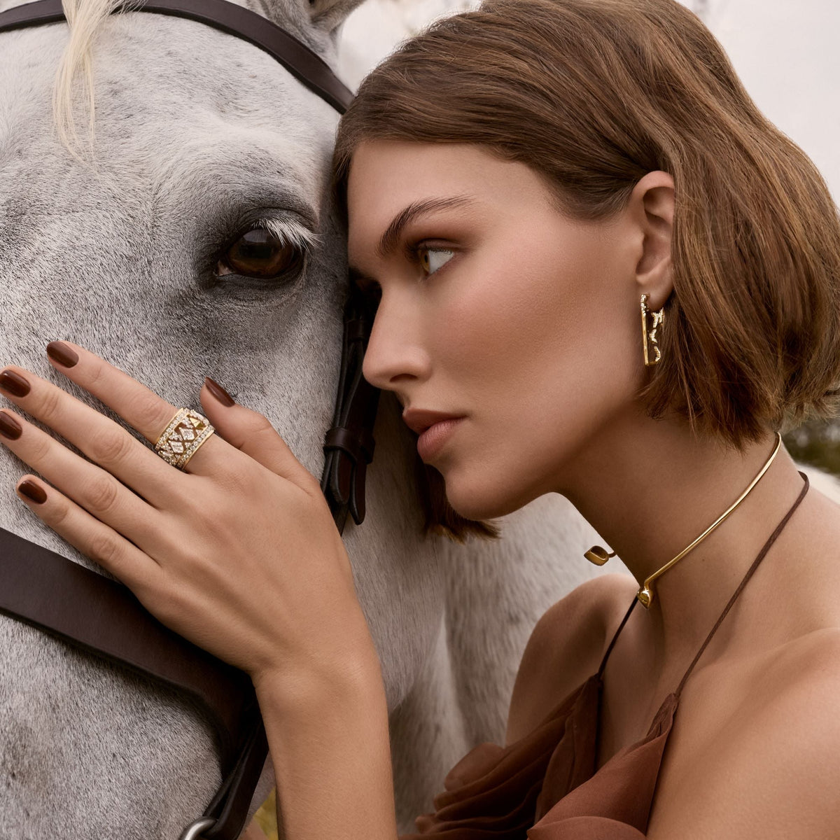 Close-up profile of a model pressing her cheek against a gray horse, wearing the assembled 14k yellow gold two-piece diamond puzzle ring on her index finger, the large diamond rectangle hoop earrings with SAV logo, and a gold collar necklace with cord.